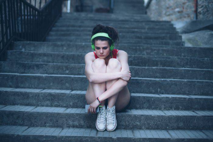 Sad girl with headphones sitting on stairs, arms around legs. Stairs, fence and stone walls on background.