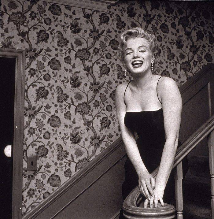 American actress Marilyn Monroe (1926 - 1962) stands in a staircase alongside a wall with a floral-motif pattern, late 1950s. She wears a black cocktail dress and dangling earrings. (Photo by Hulton Archive/Getty Images)